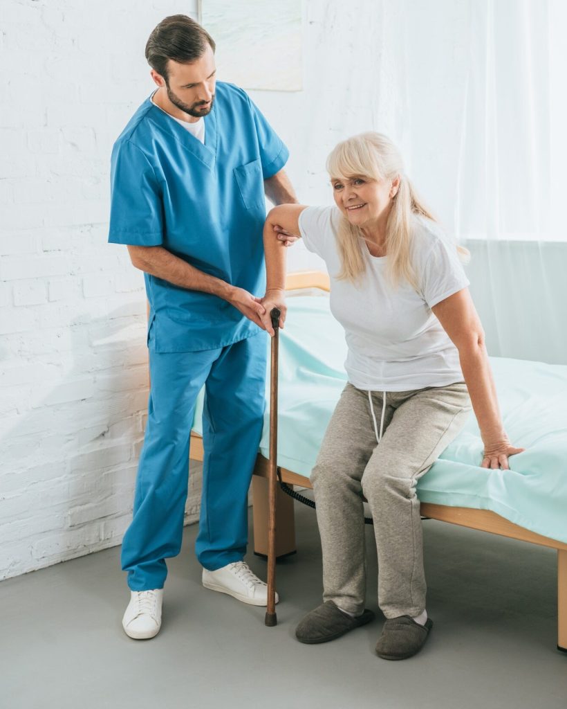 A male home care nurse helps a senior woman with a walking stick cane inside her home.