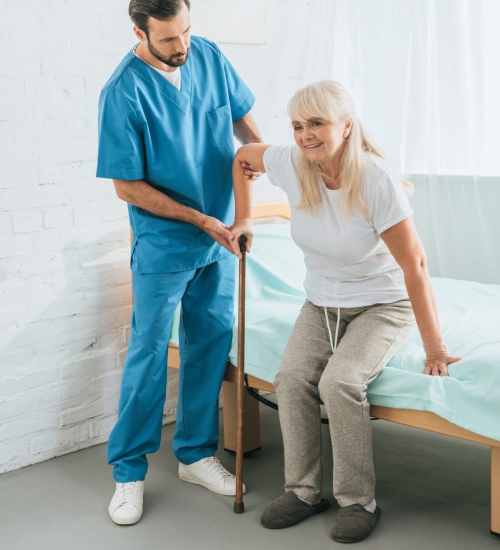 A male home care nurse helps a senior woman with a walking stick cane inside her home.