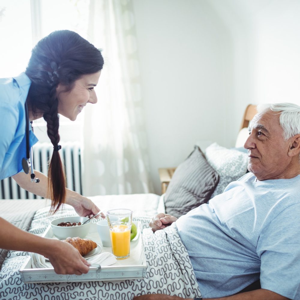 A female home care nurse serves breakfast to a senior male in the comfort of his home.