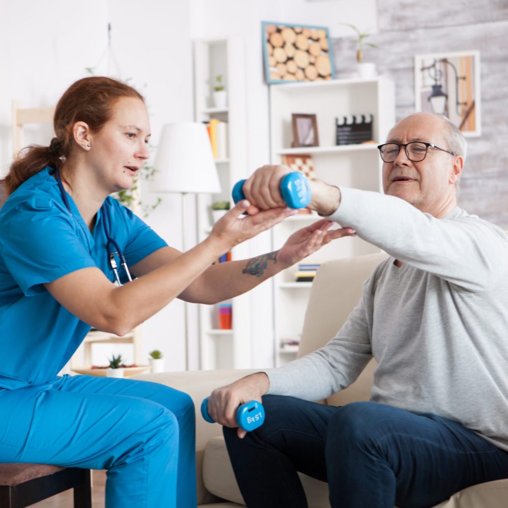 Senior man in his home doing physical therapy with help from a home care nurse using dumbbells.